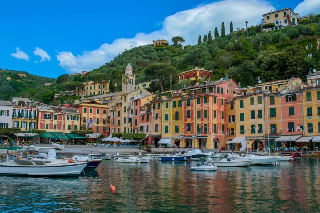 Small pleasure boats dot the harbor in Portofino, Italy, with brightly-painted buildings lining the town square beyond.