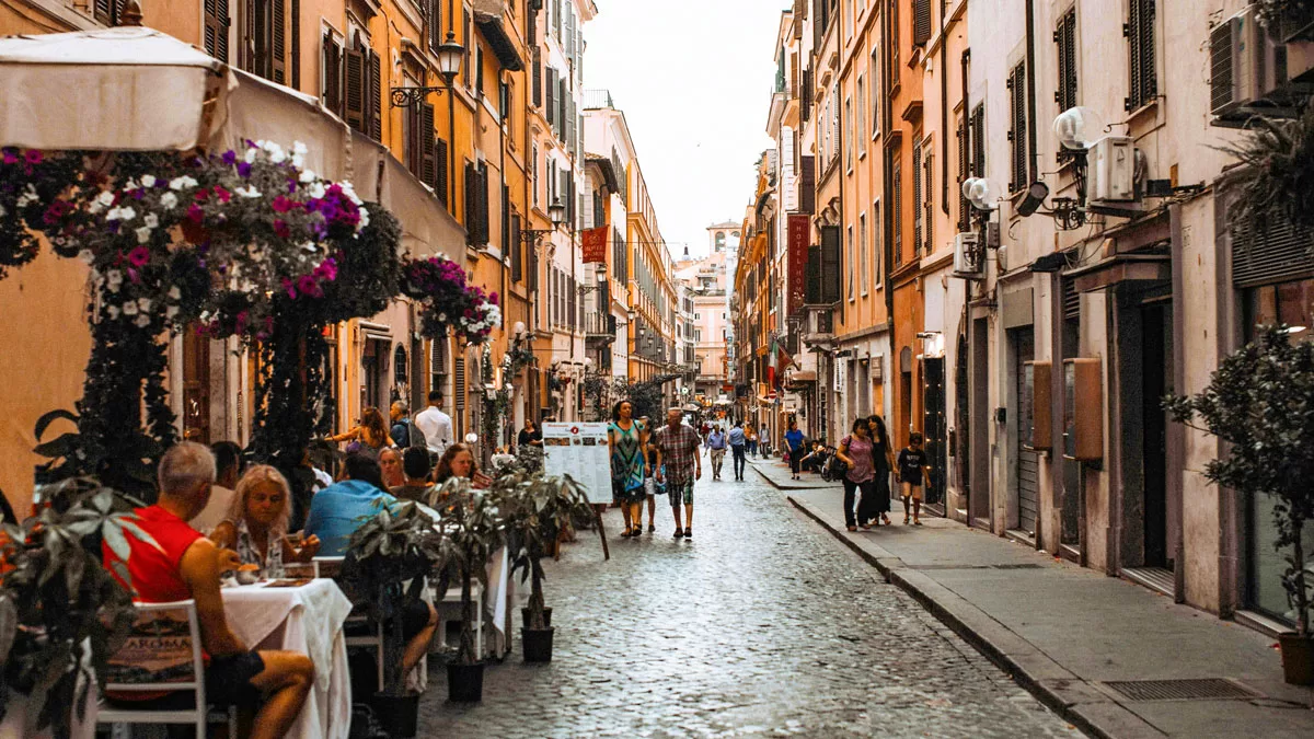 Diners sit at al fresco tables along a cobblestoned street in Rome, while pedestrians amble past.