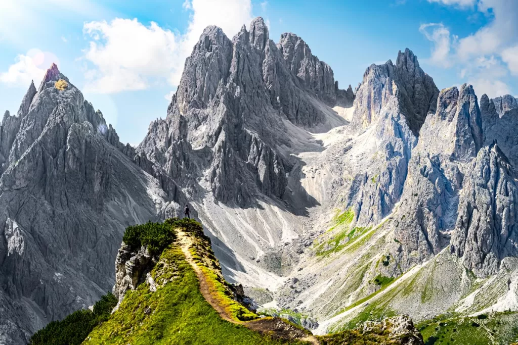 A solitary hiker stands on the narrow grassy outcropping of Cadini di Misurina, a dramatic viewpoint overlooking the Italian Dolomites.