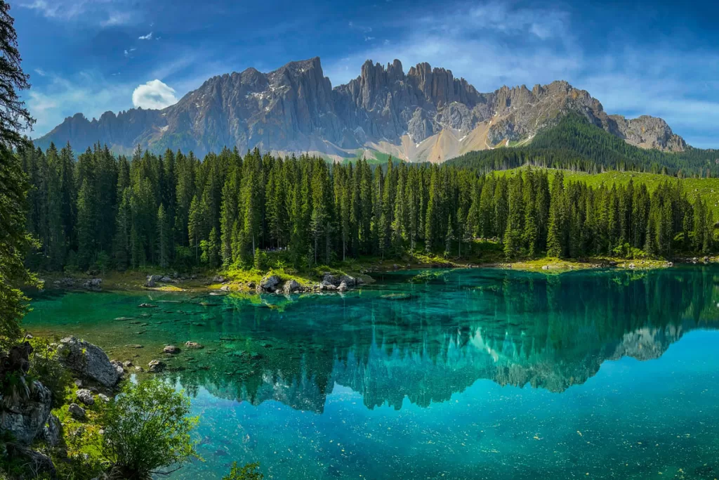 The tall, jagged peaks of Italy's Dolomite mountains are reflected in the turquoise waters of Lago di Carezza on a sunny day.