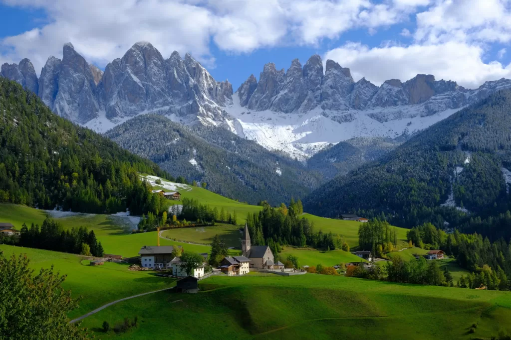 The small village church of Santa Maddalena in South Tyrol, Italy, sits in a green valley, contrasted against the stark mountain peaks of the Dolomites behind.