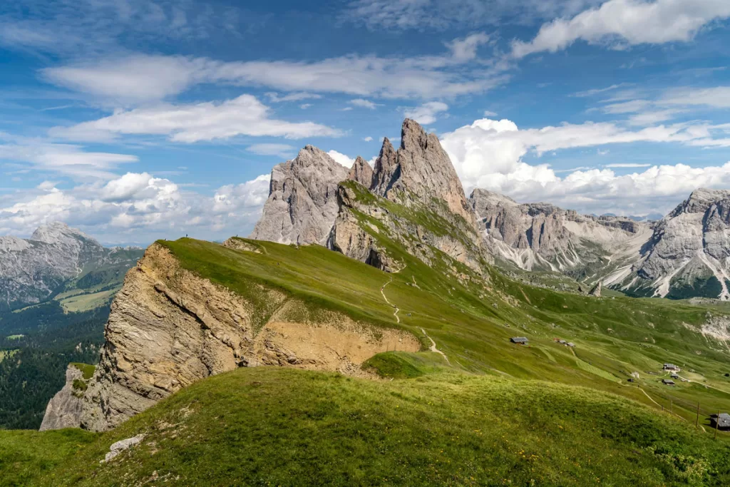 The grassy plateau of the Seceda Ridgeline slopes downward in front of tall mountain peaks. The ridge is one of Italy's most popular Dolomites photo spots.