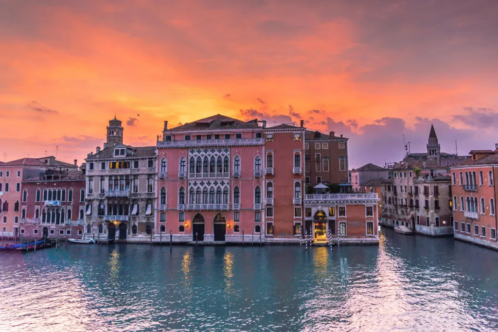 Sunset over Venice, Italy illuminates buildings along the Grand Canal in warm tones as the evening lights come on.