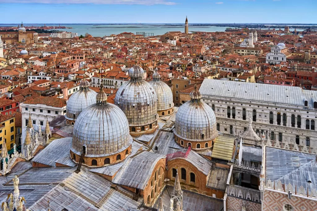 A bird's eye view of the red tiled rooftops of Venice, stretching out to the blue waters of the Venice Lagoon in the far distance.