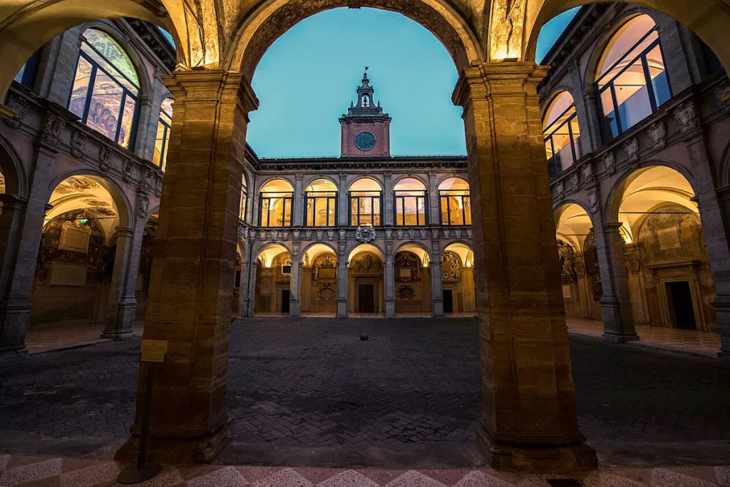 Illuminated arches frame the central courtyard of the University of Bologna's Archiginnasio at dusk.