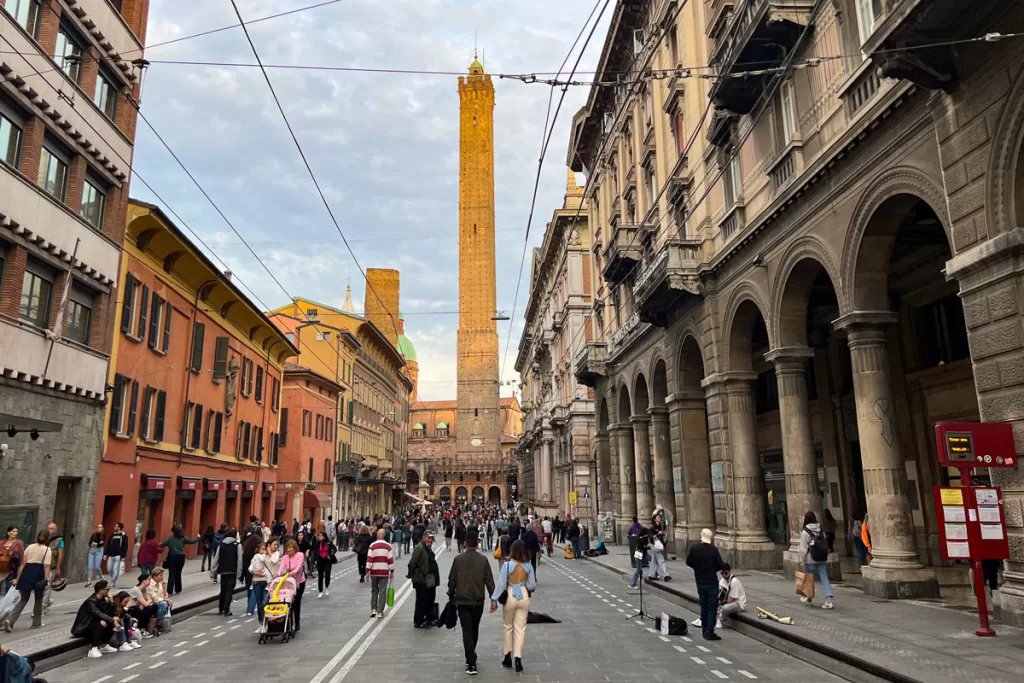 Evening light shines on the Asinelli Tower above a crowded pedestrian street in Bologna, Italy.