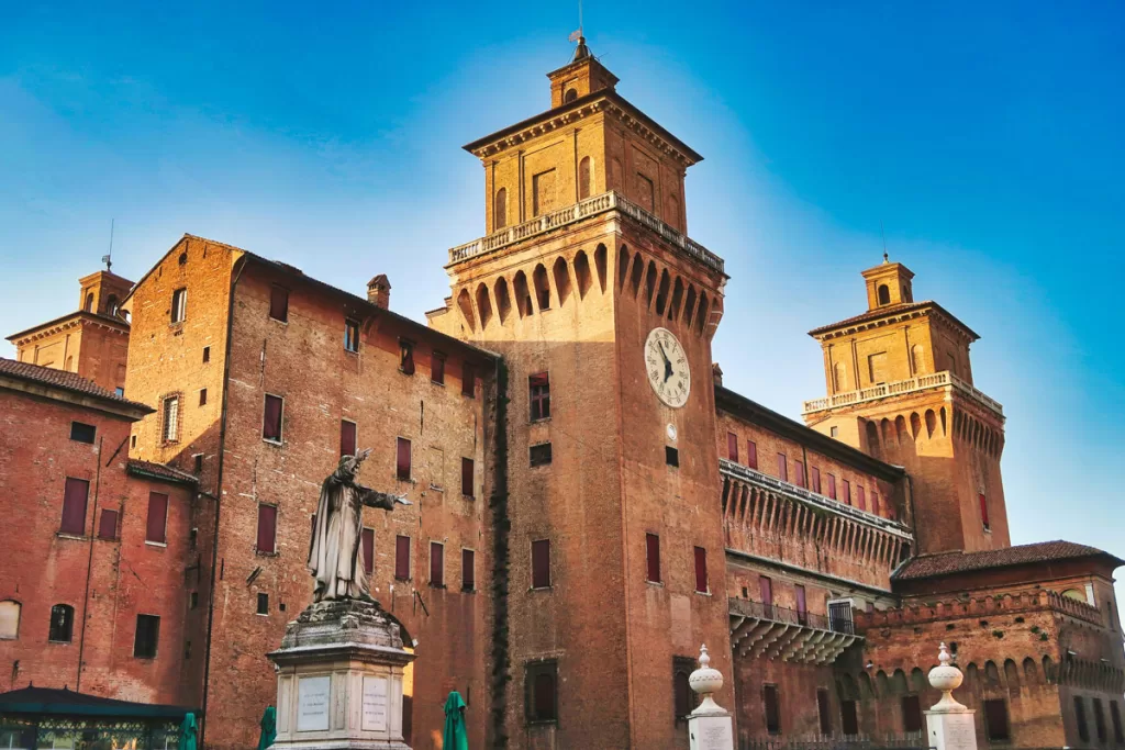A stone statue of a man with outreached arms stands in front of the dominant Castello Estense in Ferrara, Italy