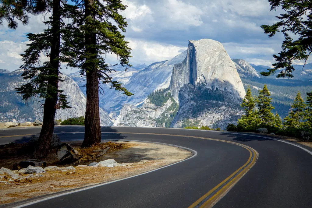 A two-lane road winds sharply around a mountain bend, looking out to Half Dome in Yosemite National Park. The vista is an iconic image, and one of the best photo spots in Yosemite.