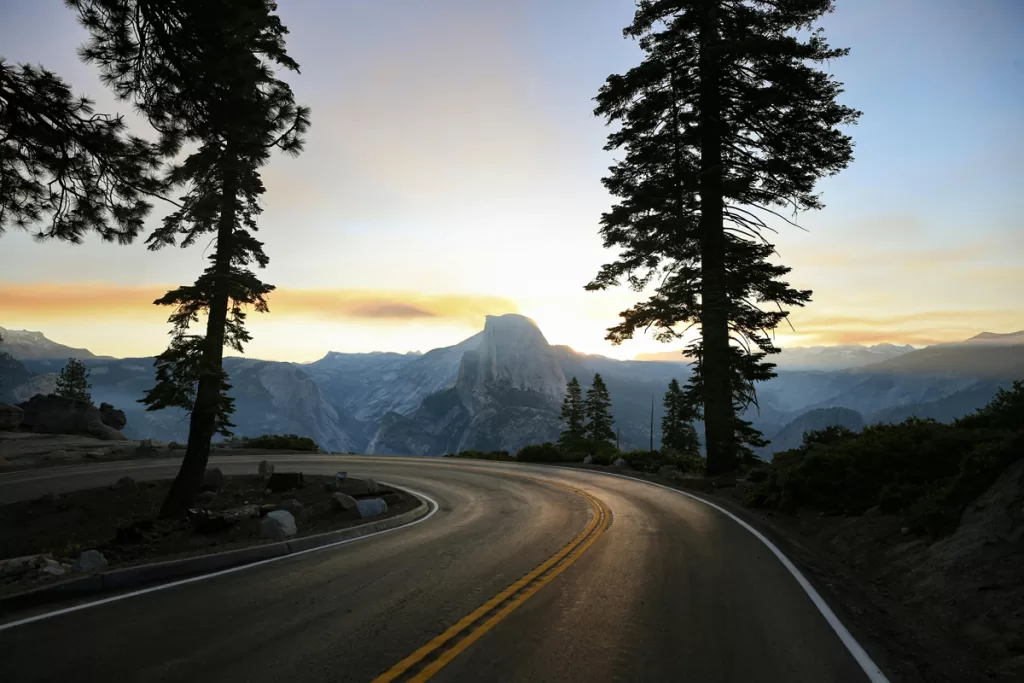 A two-lane road turns sharply on a mountain edge, overlooking Half Dome at dusk in Yosemite National Park.