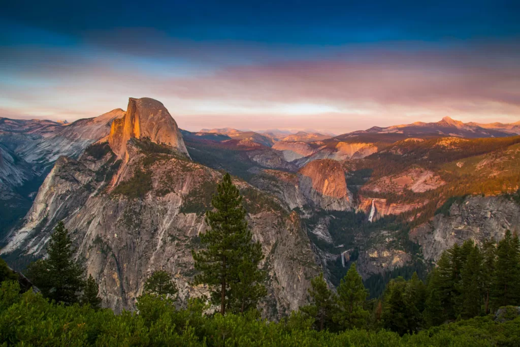 Sunset at Glacier Point in Yosemite National Park illuminates Half Dome and Vernal Falls in warm pink and orange tones.