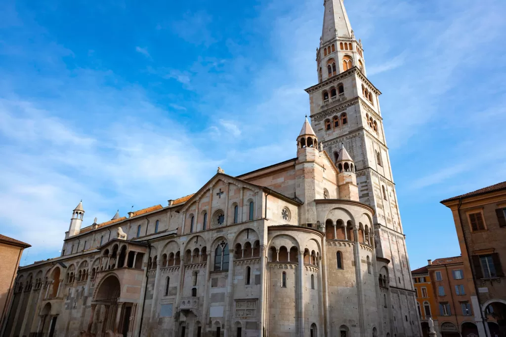 The stone facade of the medieval Modena Cathedral rises against a blue sky backdrop.