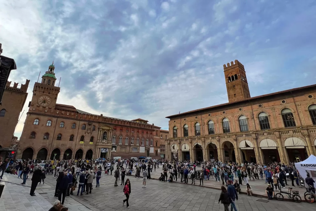 Pedestrians crowd the sidewalks of Piazza Maggiore in Bologna, Italy, surrounded by the city's iconic medieval brick buildings.