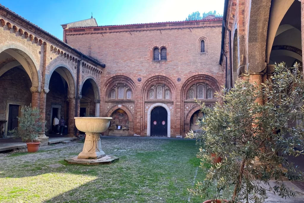 A stone fountain stands in the walled central courtyard of the Basilica di Santo Stefano in Bologna, Italy, flanked by potted olive trees.