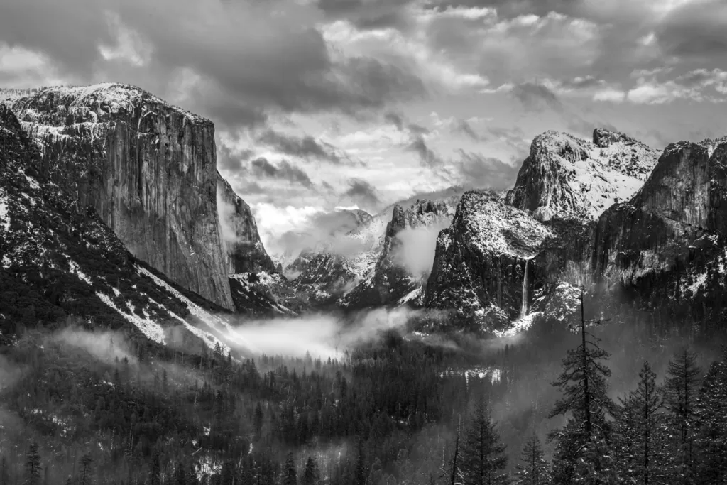 A black and white photograph of Yosemite Valley after a winter storm from the Tunnel View overlook, with mist shrouding the valley and El Capitan and Bridalveil Falls dusted with snow.