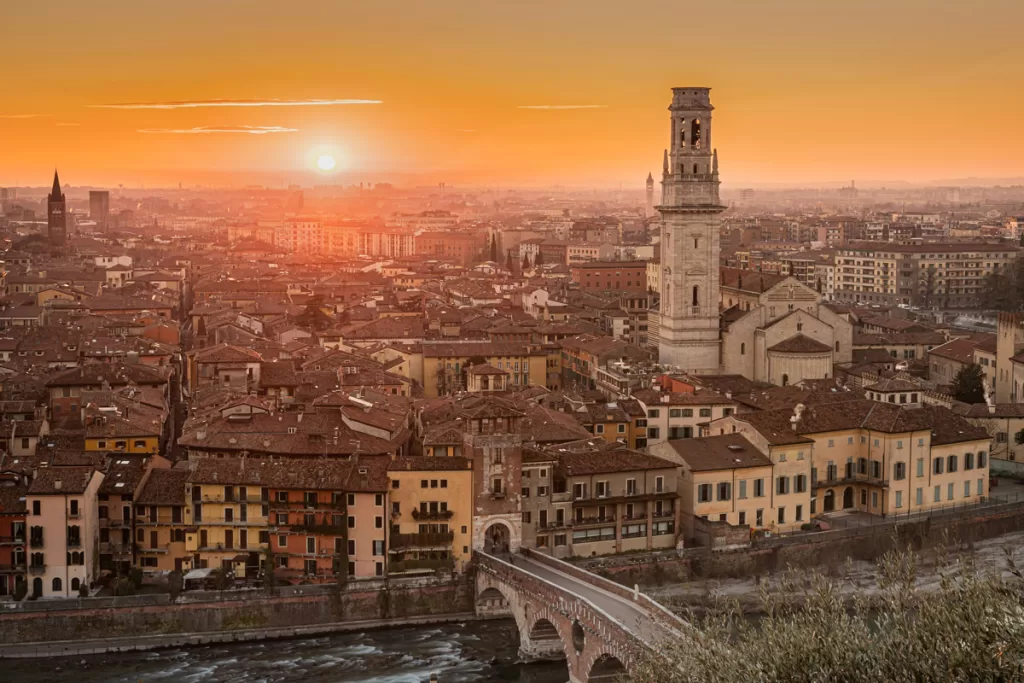 Sunset casts a warm glow over the rooftops of Verona, Italy, with the cathedral bell tower rising prominently in the foreground.