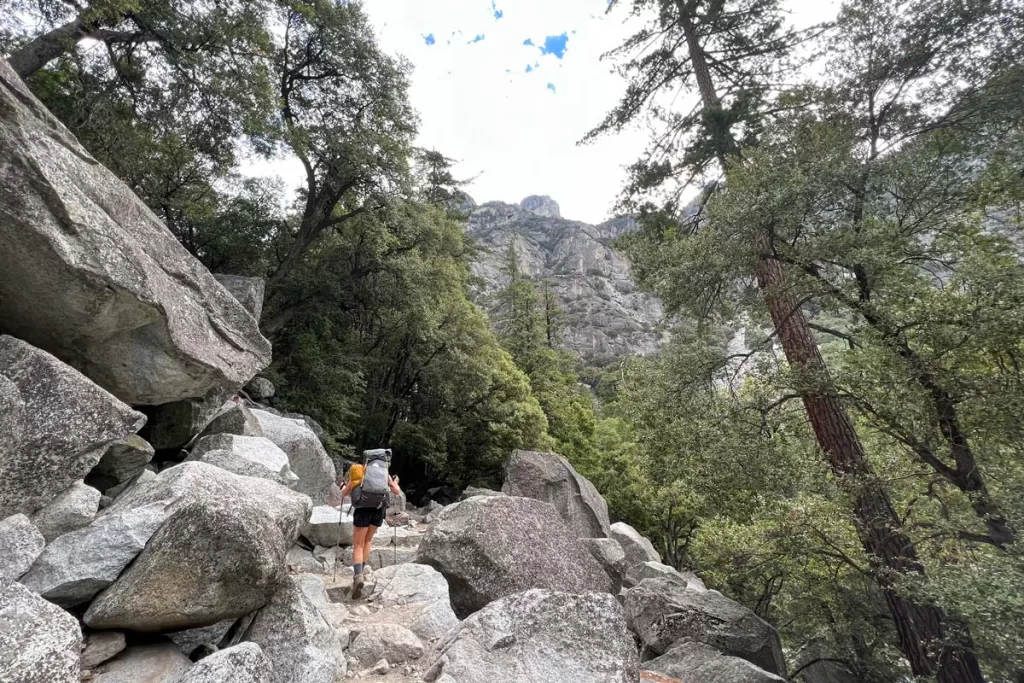 A female hiker climbs granite rock steps in a forested stretch of the Rae Lakes Loop in Kings Canyon National Park.