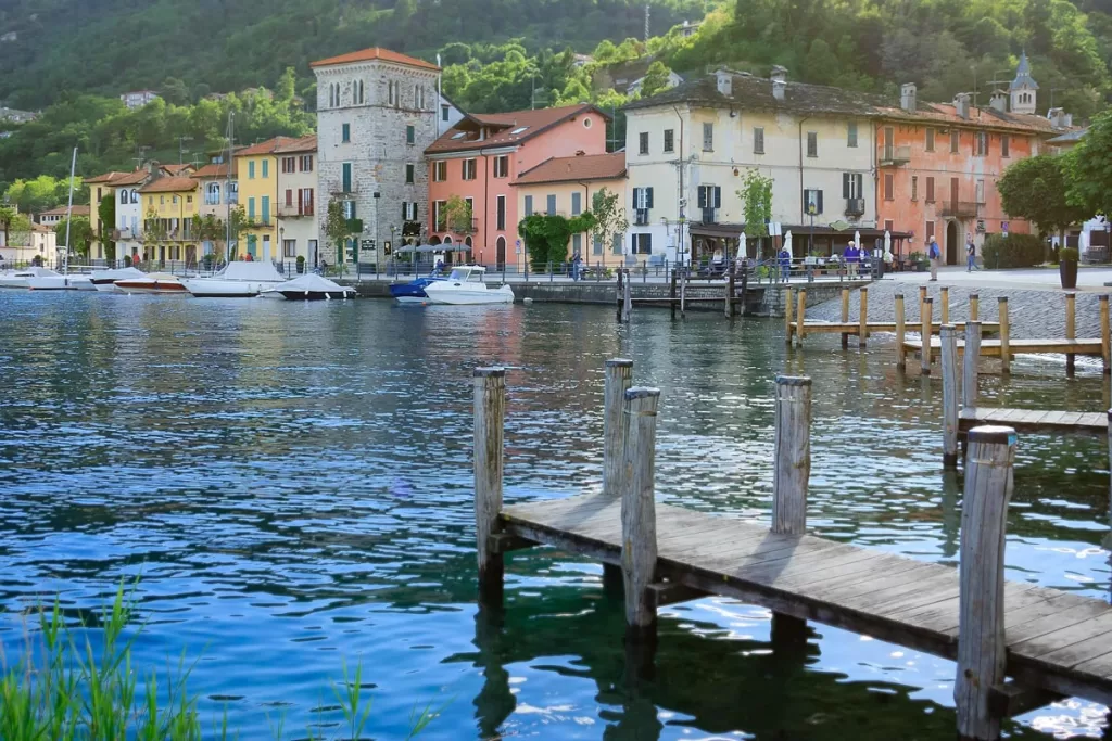A wooden dock juts out over the waters of Lake Orta in Lombardy, Italy, with pastel buildings lining the shoreline in the background. 