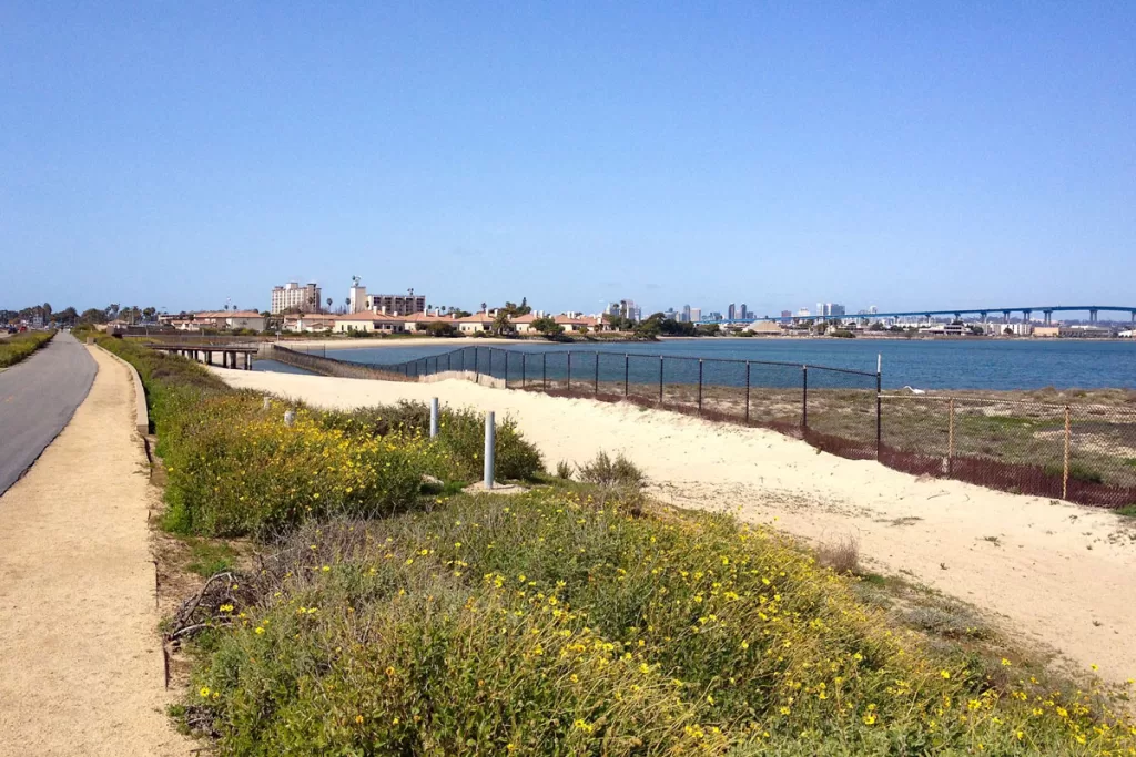 A paved bike path hugs the sandy shoreline of San Diego Bay along the Bayshore Bikepath, with the Coronado Bridge visible in the distance.