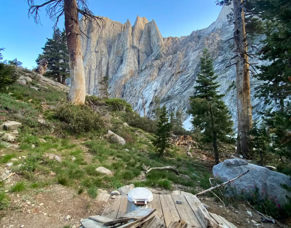 A solitary pit toilet sits on a ridge in California's Sierra Nevada backcountry, overlooking tall granite peaks across the valley.