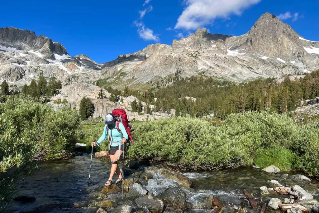 A female hiker crosses a small creek, surrounded by scrub brush, in California's Sierra Nevada mountains. Waterproof sandals are strongly encouraged when making a backpacking packing list.