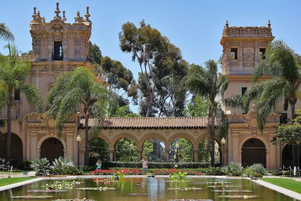 Tourists stand by a reflecting pond in San Diego's Balboa Park neighborhood, with the city's iconic Spanish Colonial architecture in the background.