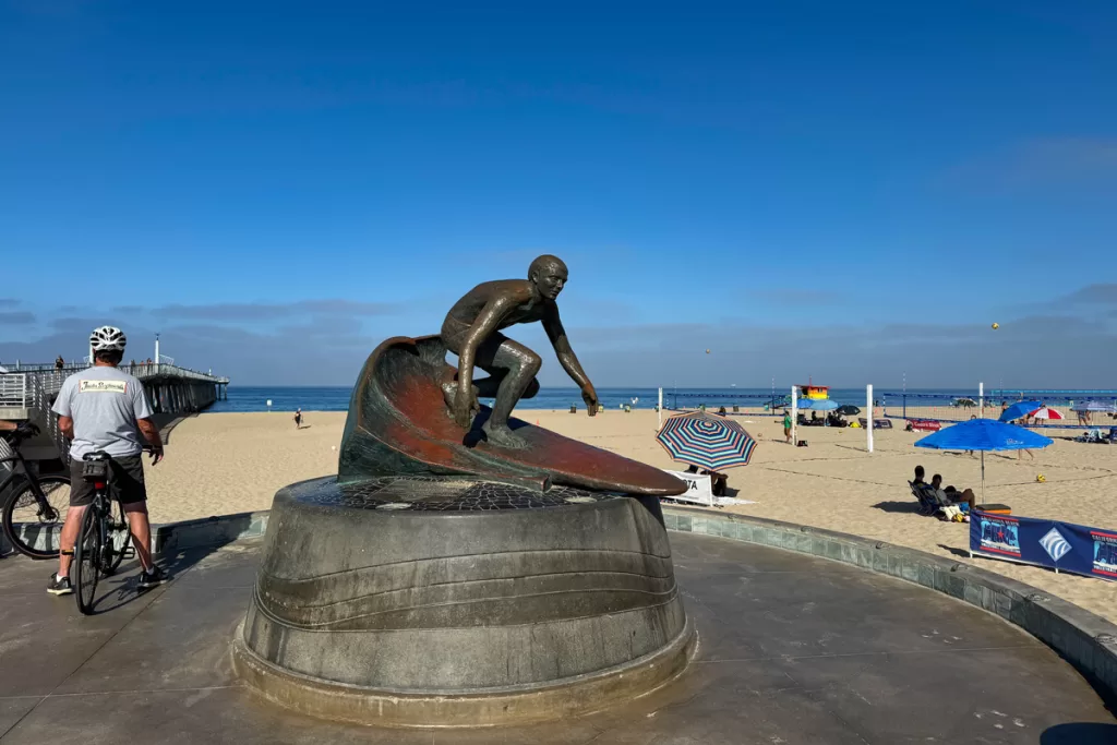 The iconic Tim Kelly surf statue against a backdrop of beach umbrellas and volleyball courts in Hermosa Beach, California.