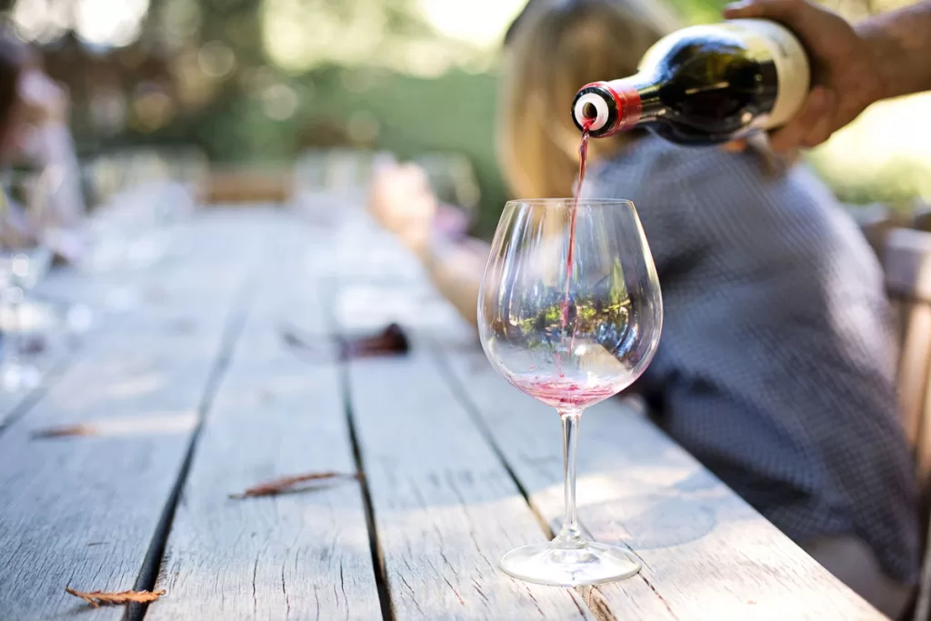 A tasting is poured into a wine glass on an outdoor wooden table.