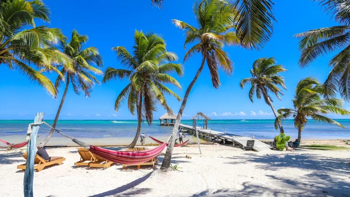A beach scene in Ambergris Caye, Belize, with wooden lounge chairs, a striped hammock, and wooden dock. Ambergris Caye is the top destination for most Belize itinerary guides.