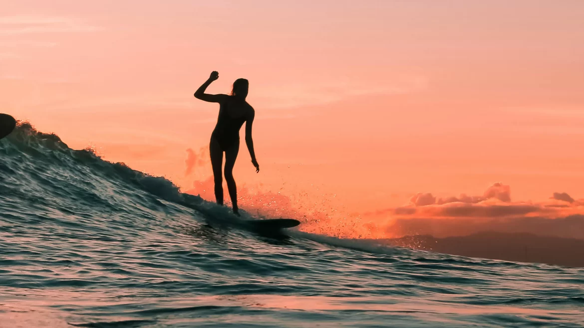 The silhouette of a woman surfing at sunset is backlit against a pink sky