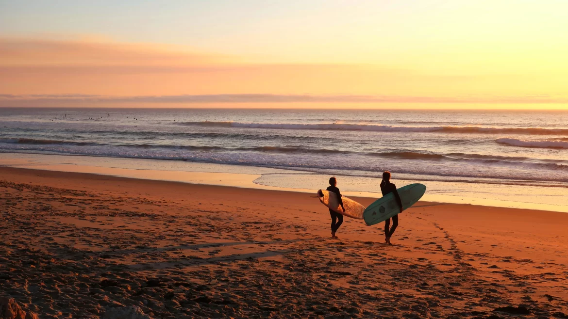 Two surfers stand on shore overlooking a beach at sunset.